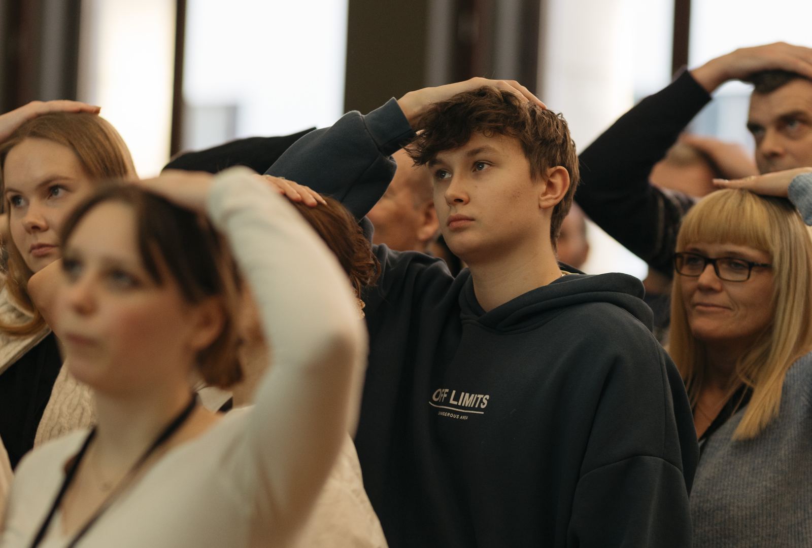 Young people and adults stand together with a hand on their heads during an exercise.